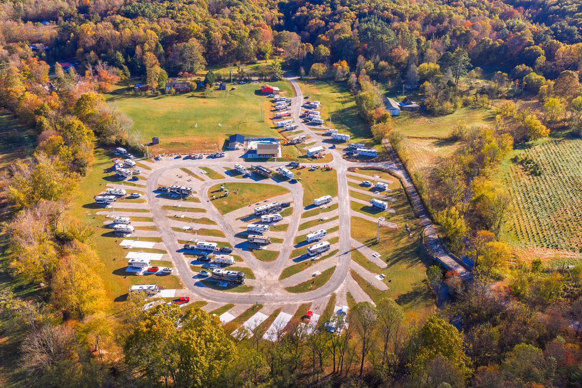 A high-angle view of an RV park set in a rural landscape, surrounded by autumn foliage with cars and trailers parked in designated spots.